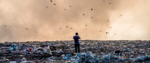 Image of a person standing in the middle of a dump/landfill. the sky is sepia and there are birds flying around. Image by Bakhrom Tursunov from Pixabay.